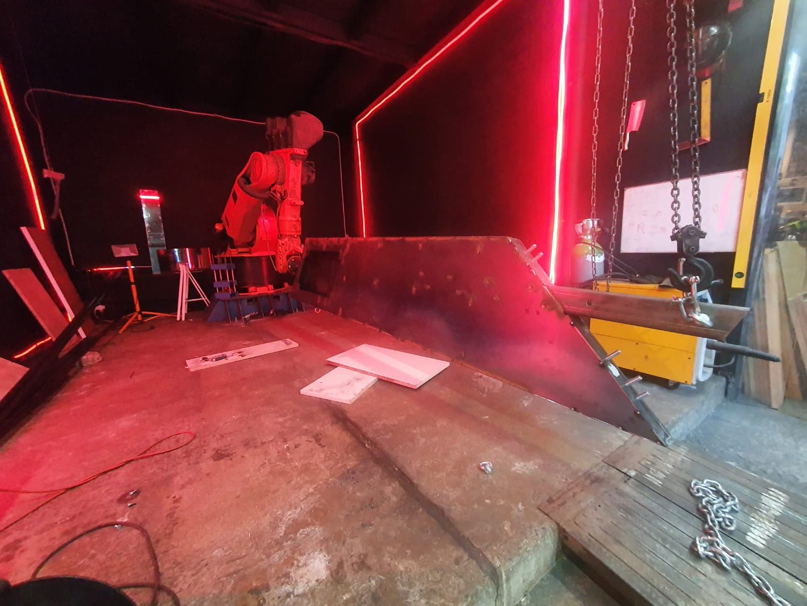 A workshop under red light: an industrial robot arm beside one leg of the swing frame in bare steel, lifting chains at right.