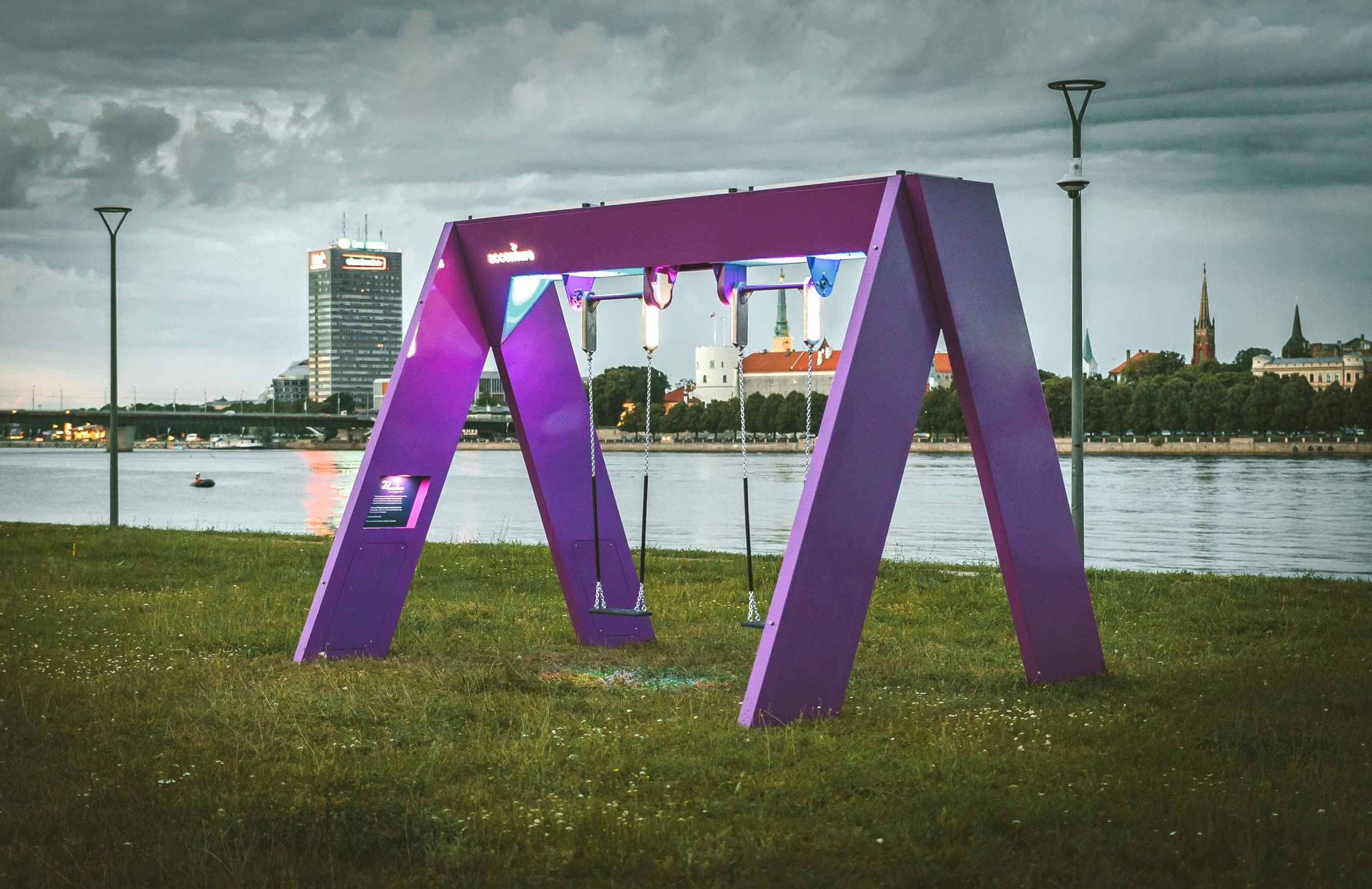 The purple swing set on a riverbank lawn at dusk, Riga Castle and the old town across the Daugava behind it.