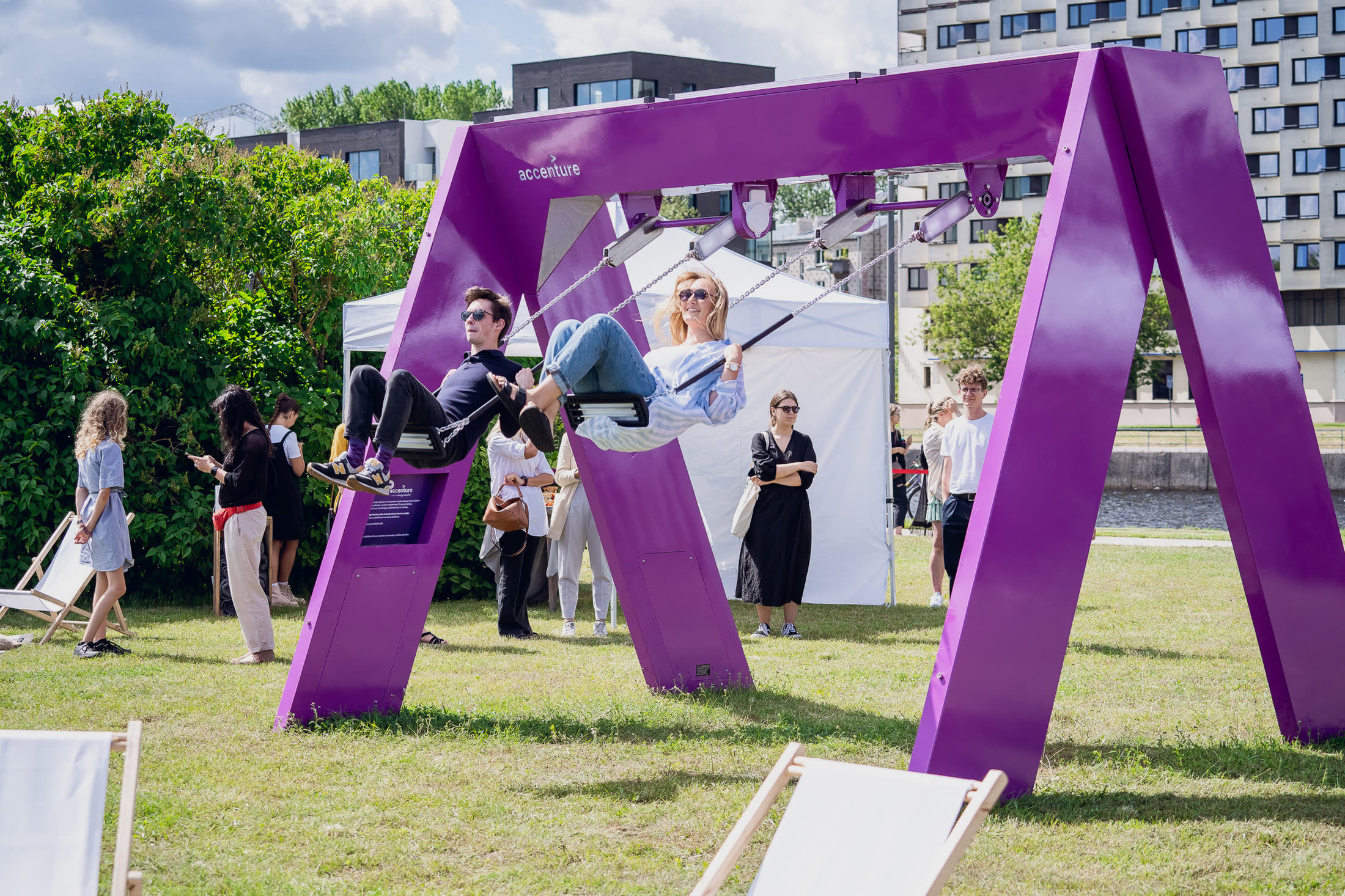 A man and a woman in sunglasses swinging together and laughing, an event tent behind them.
