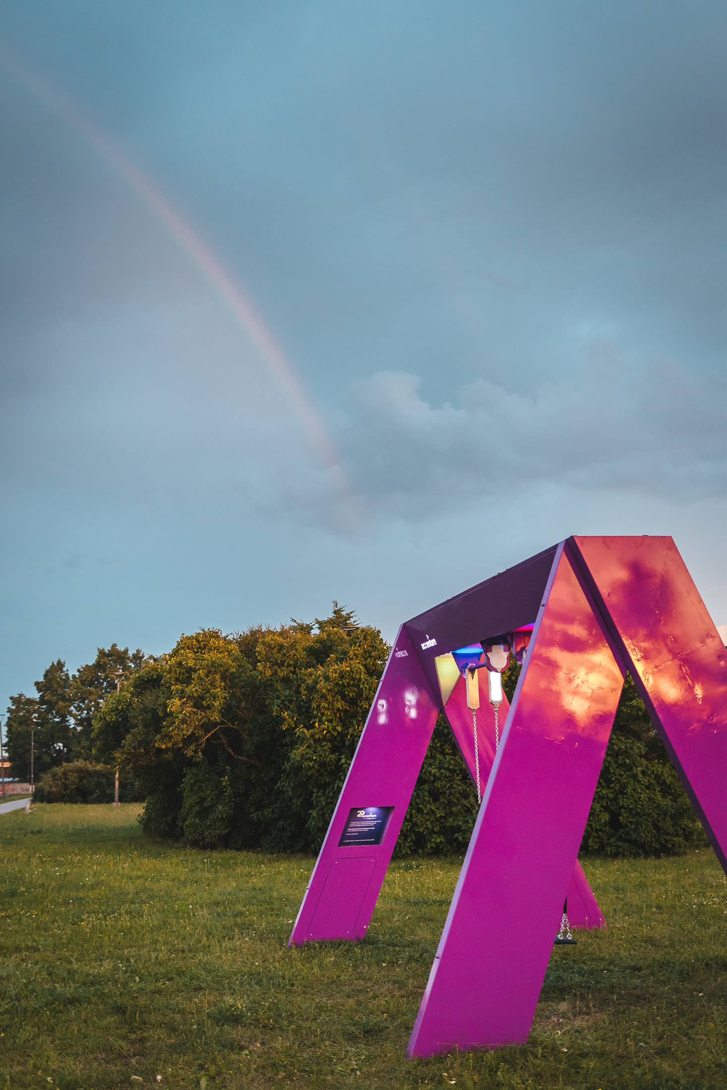 The swing structure from a low angle at dusk, a rainbow arcing across the grey sky above it.