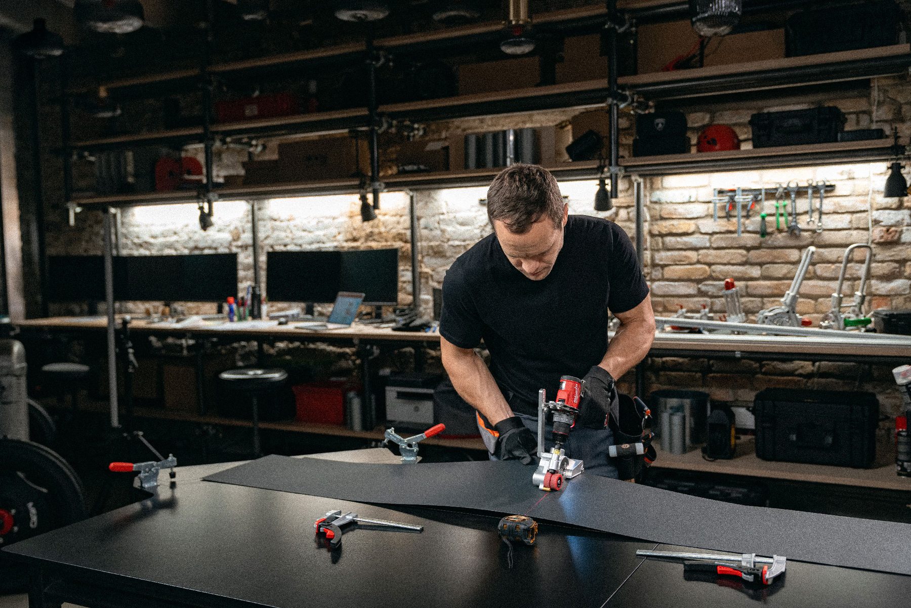 A man in a black T-shirt works a seaming tool along a dark metal sheet in a brick workshop.