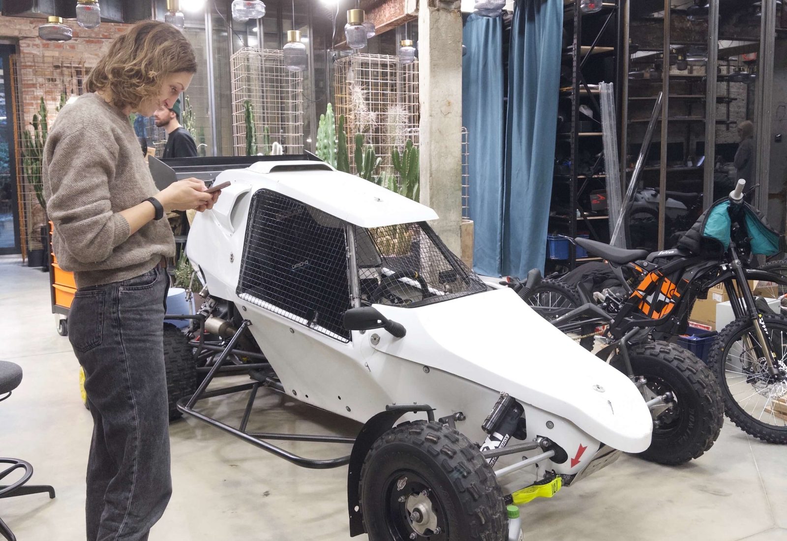 A woman checks her phone beside a white off-road buggy with a roll cage, parked inside the workshop next to a motorbike.