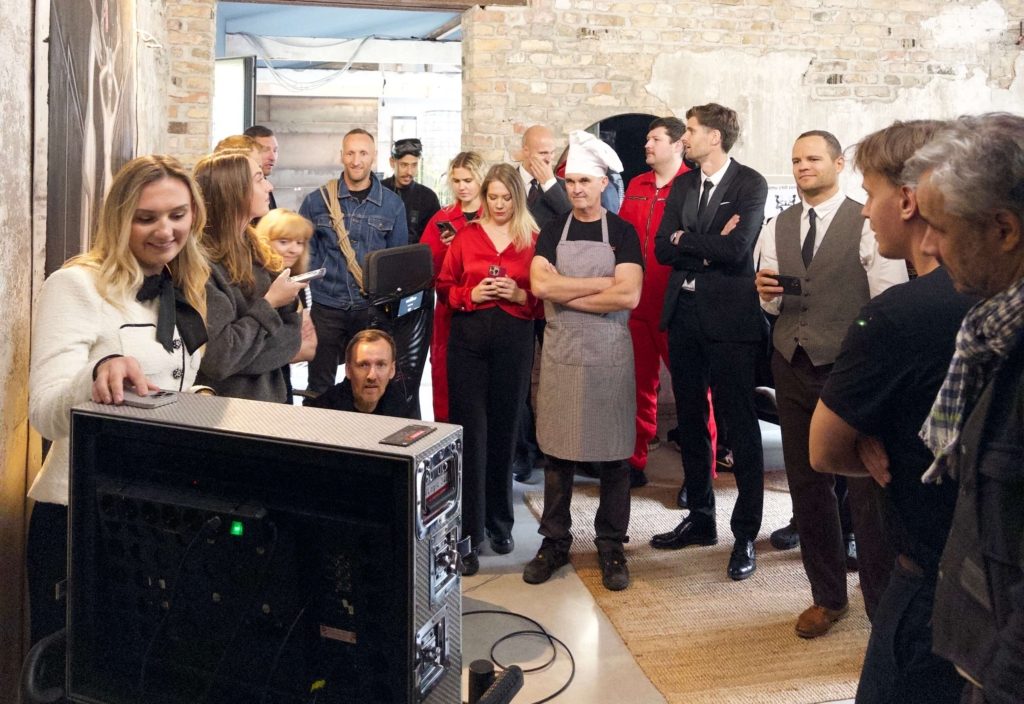 About fifteen people packed into a brick room, a chef in whites among them, all watching playback off-screen.