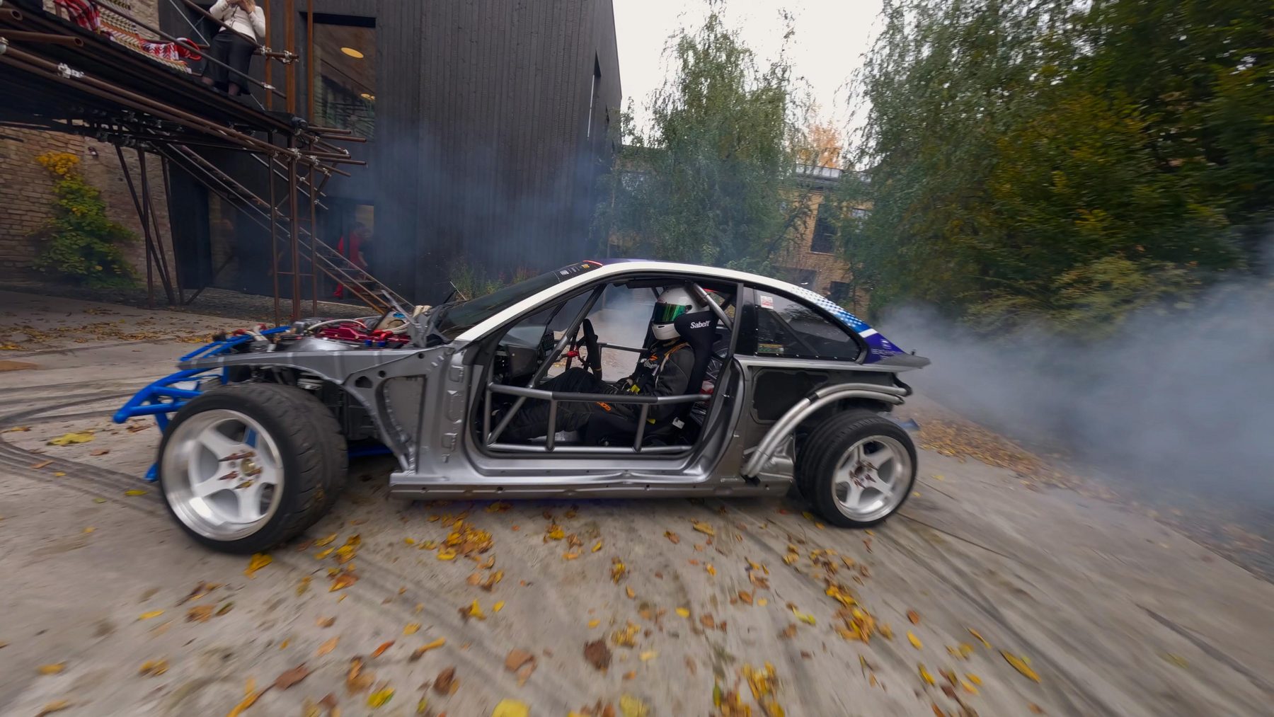 A car stripped to its bare frame sliding sideways through fallen leaves in an industrial yard, tyre smoke pouring off the rear wheels.