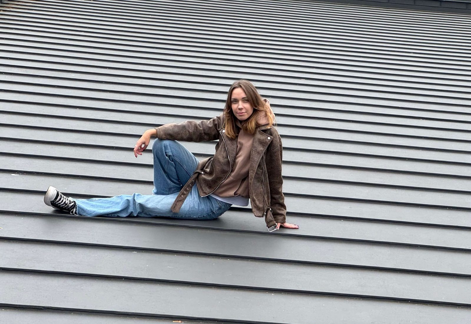 A woman in a brown jacket sits on a grey standing-seam metal roof, the seams running diagonally across the frame.