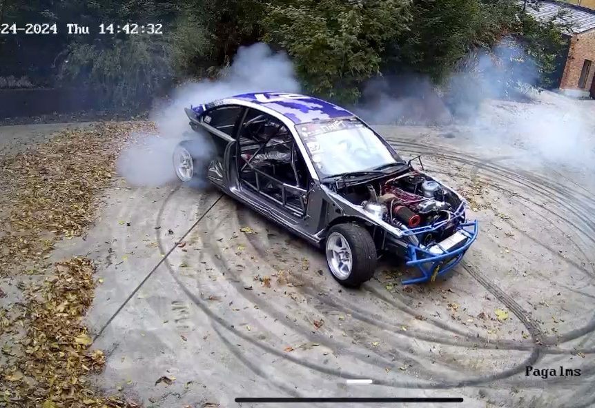 A timestamped security-camera frame of the skeletal car mid-drift in a cloud of tyre smoke.