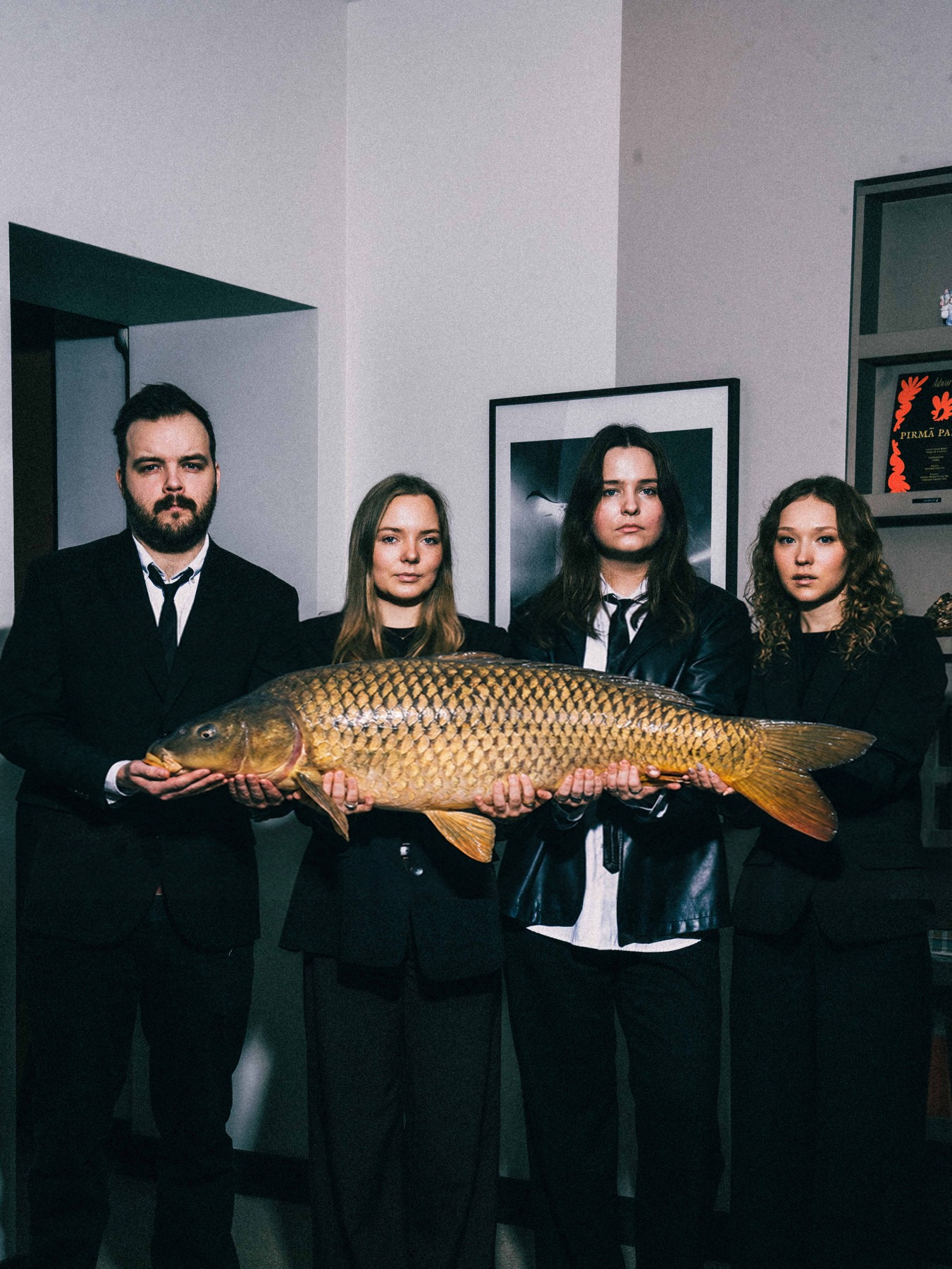 Four people in black suits stand shoulder to shoulder, deadpan, holding a very large carp across all of their hands.