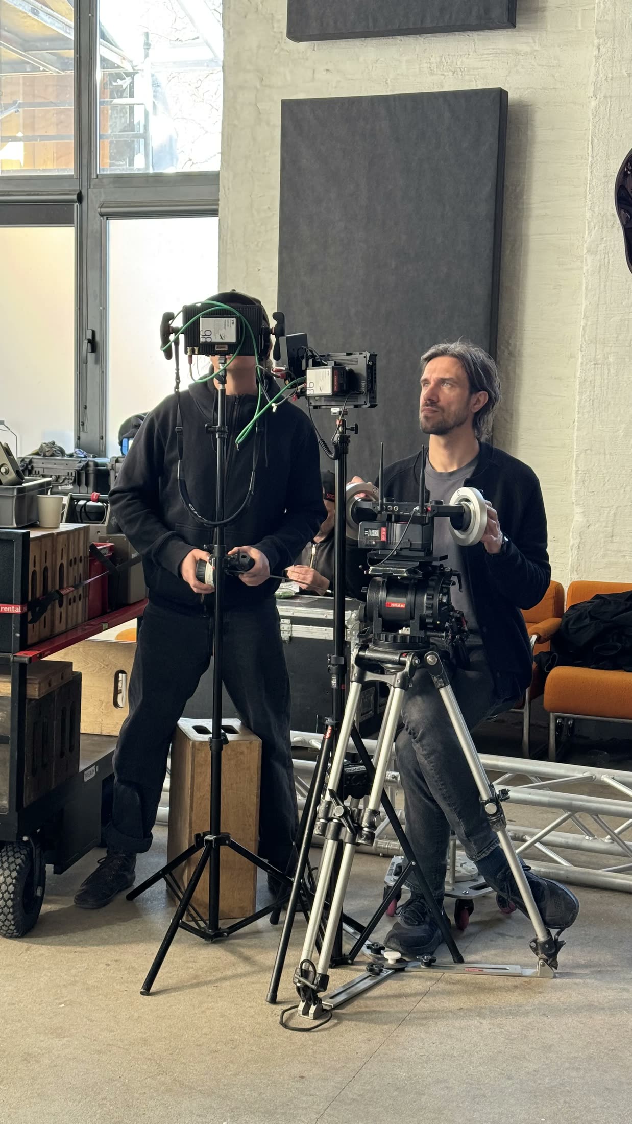 Two crew members in a daylit loft studio, one behind a monitor with a follow-focus, one behind a cinema camera, both looking up.