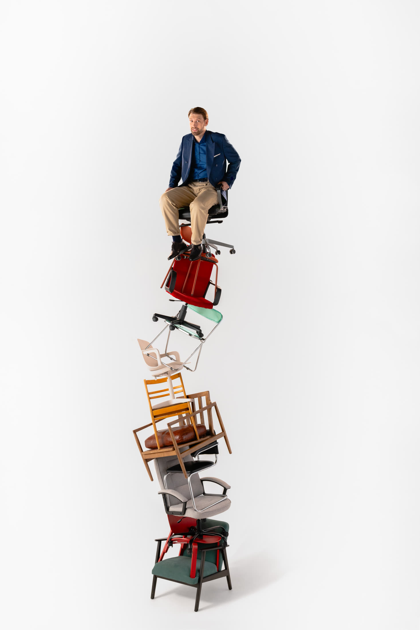 A bearded man in a navy blazer sits on an office chair at the top of a tall stack of about ten mismatched chairs, against plain white.