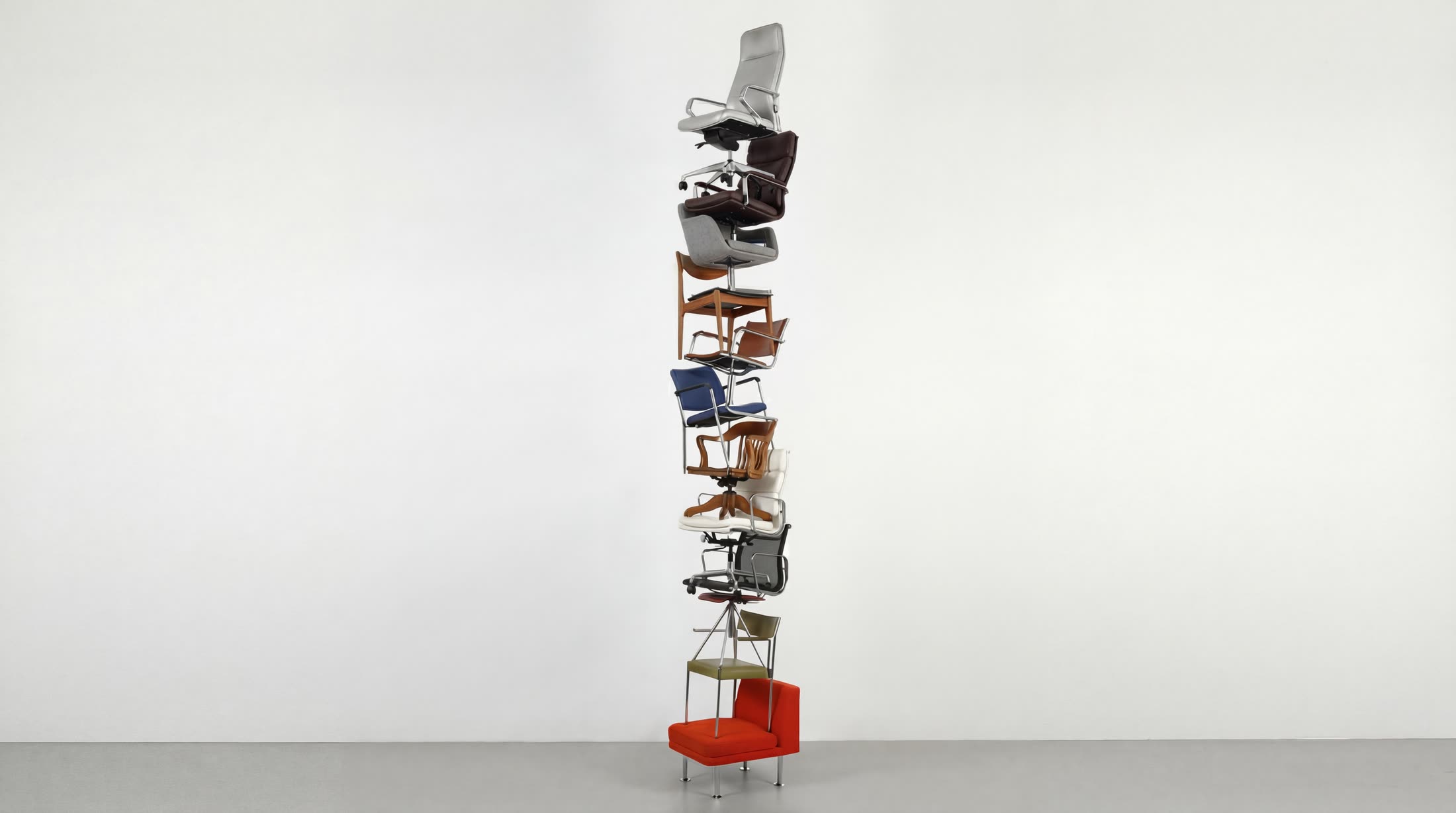 A column of about twelve stacked, interlocked chairs of different styles on a grey floor against a light-grey wall, with no person.