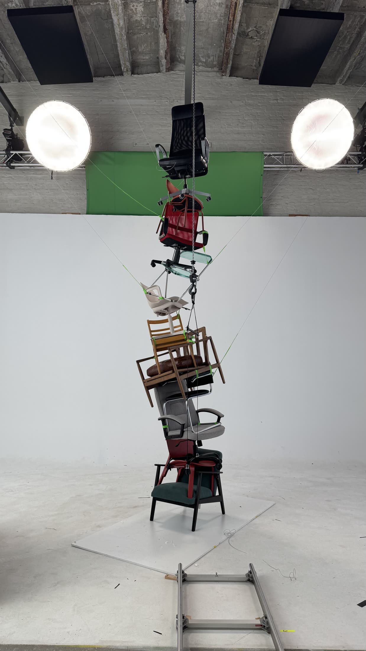 The chair stack in a studio, suspended and tied with bright green cord to a chain hoist and guy-wires, softboxes and a white cyclorama around it.