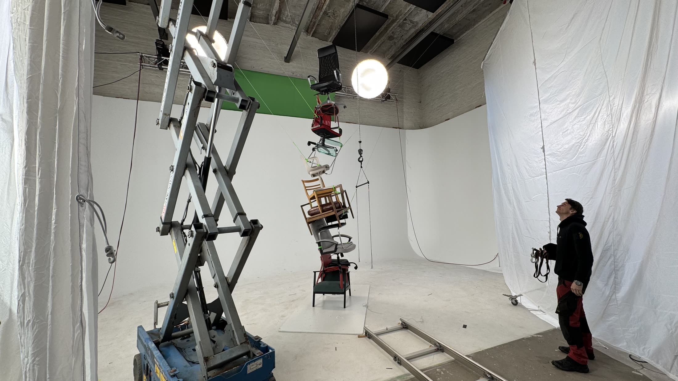Wider studio view: the rigged tower hanging mid-frame with green tie-downs, a blue scissor lift, dolly track and a crew member looking up.