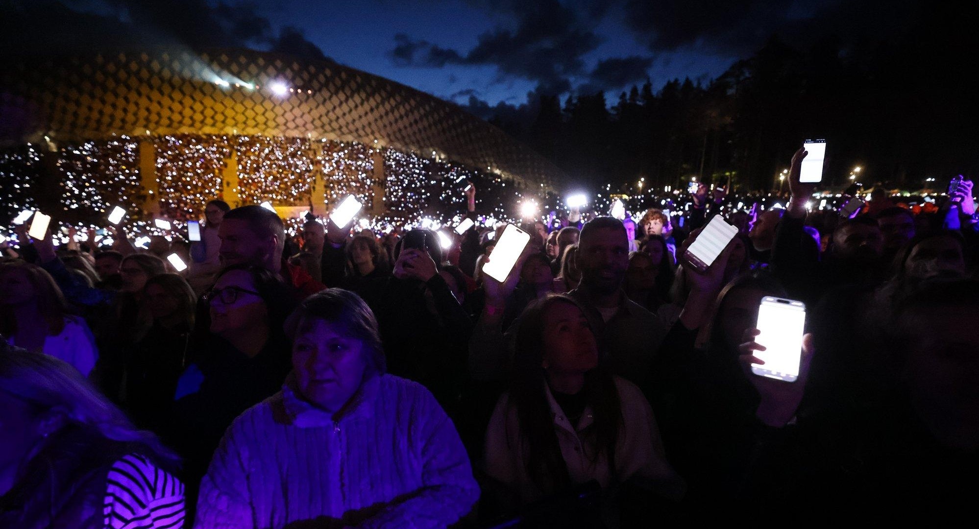 Inside the crowd at night: people holding phones overhead with white-lit screens, the stands behind dotted with thousands more.