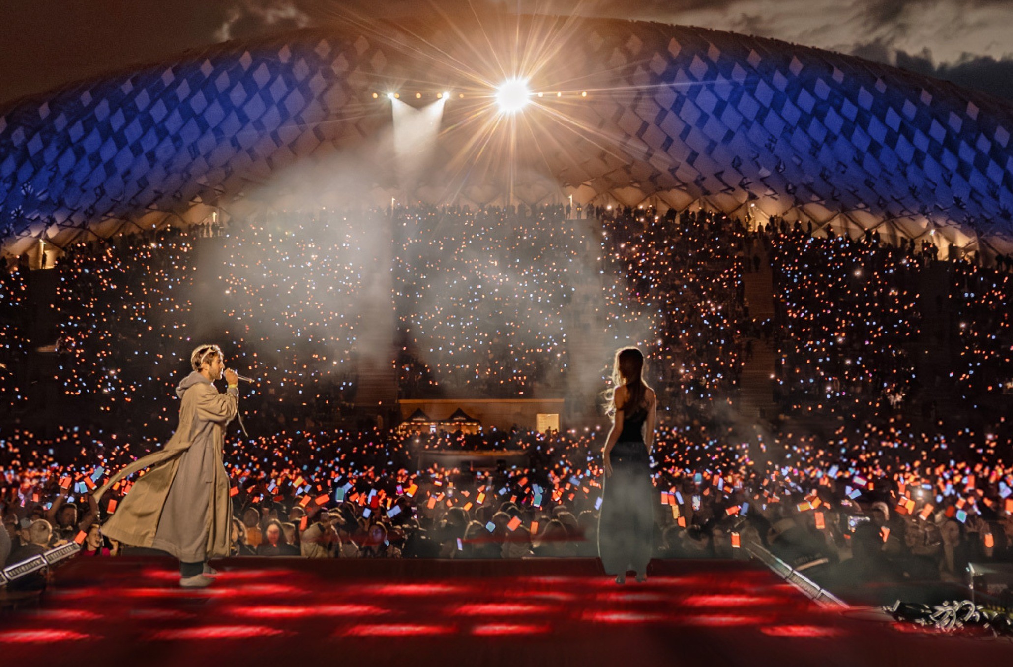 From the stage at Mežaparks at night: two singers, and behind them the whole amphitheatre filled with phone screens glowing red, orange and blue.
